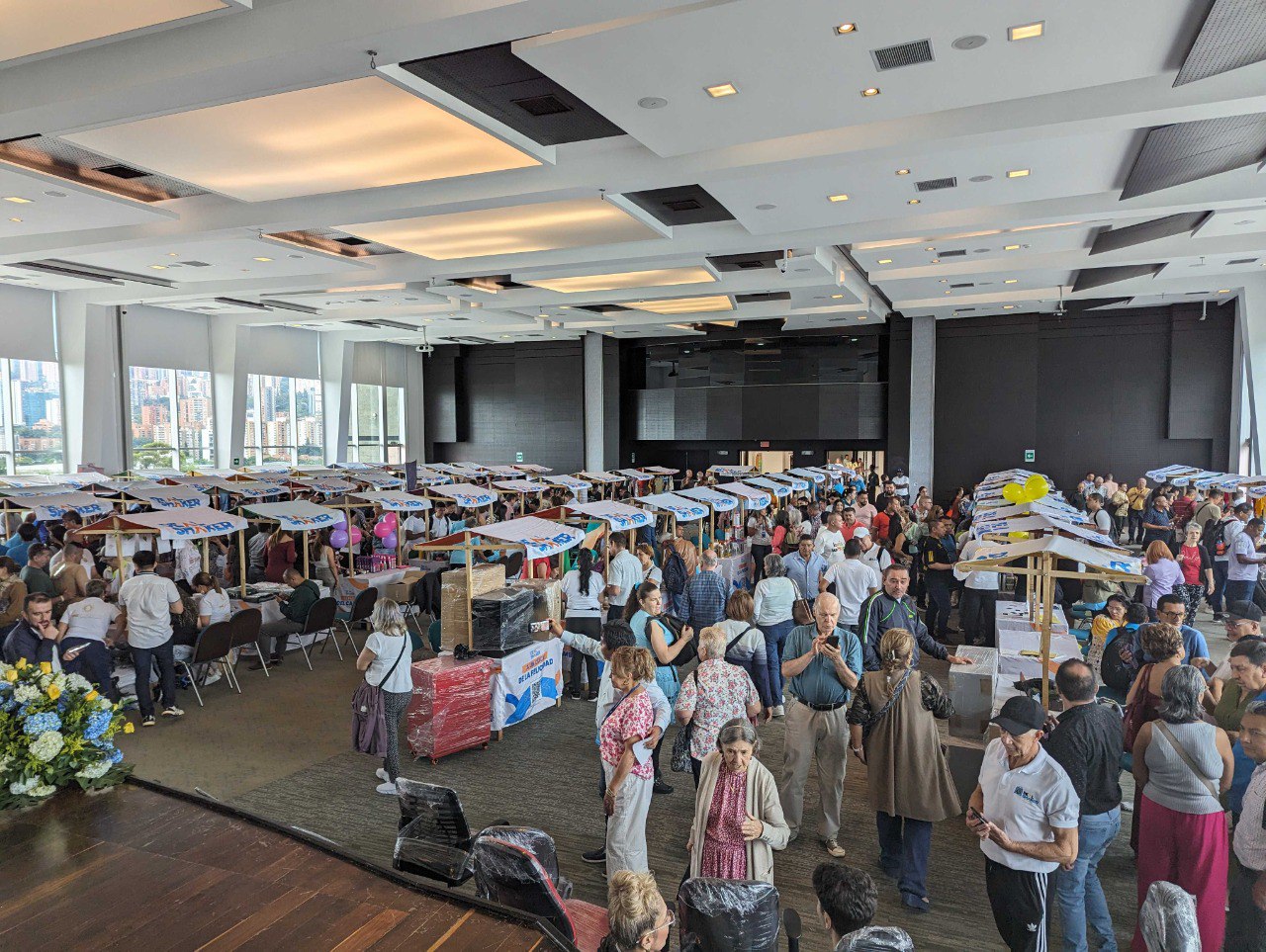 L'Import Resale Cross-border EC è un evento al coperto su larga scala in cui un gran numero di persone si alza e conversa attorno a un tavolo con vari oggetti esposti in una sala con grandi finestre e vista sulla città.